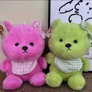Puede incluir: Se muestran dos osos de peluche, uno rosa y otro verde. Cada oso tiene un lazo, un babero con estampado de cuadros y una etiqueta. Los osos tienen ojos y narices negras. El fondo es de color neutro.