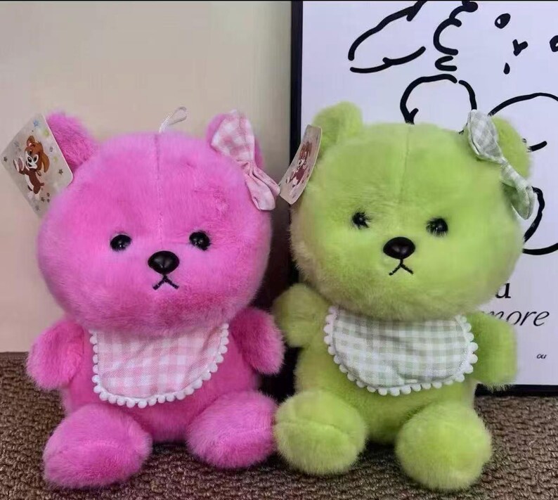 Puede incluir: Se muestran dos osos de peluche, uno rosa y otro verde. Cada oso tiene un lazo, un babero con estampado de cuadros y una etiqueta. Los osos tienen ojos y narices negras. El fondo es de color neutro.