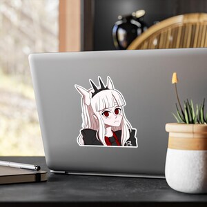May include: A sticker featuring an anime-style character with long white hair, red eyes, and a black crown. The character wears a black jacket over a red top. The sticker is affixed to a gray laptop.