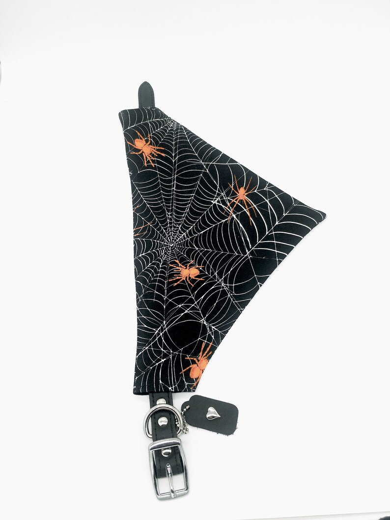 Op de afbeelding: Zwarte hondenbandana met een wit spinnenwebpatroon en oranje spinnen. De bandana is bevestigd aan een zwarte leren halsband met een zilveren gesp en een klein label.