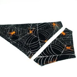 Op de afbeelding: Zwart-witte Halloween-bandana en bijpassend accessoire voor huisdieren. De stoffen items hebben een spinnenwebontwerp met oranje spinnen. De bandana en het accessoire worden op een witte achtergrond weergegeven.