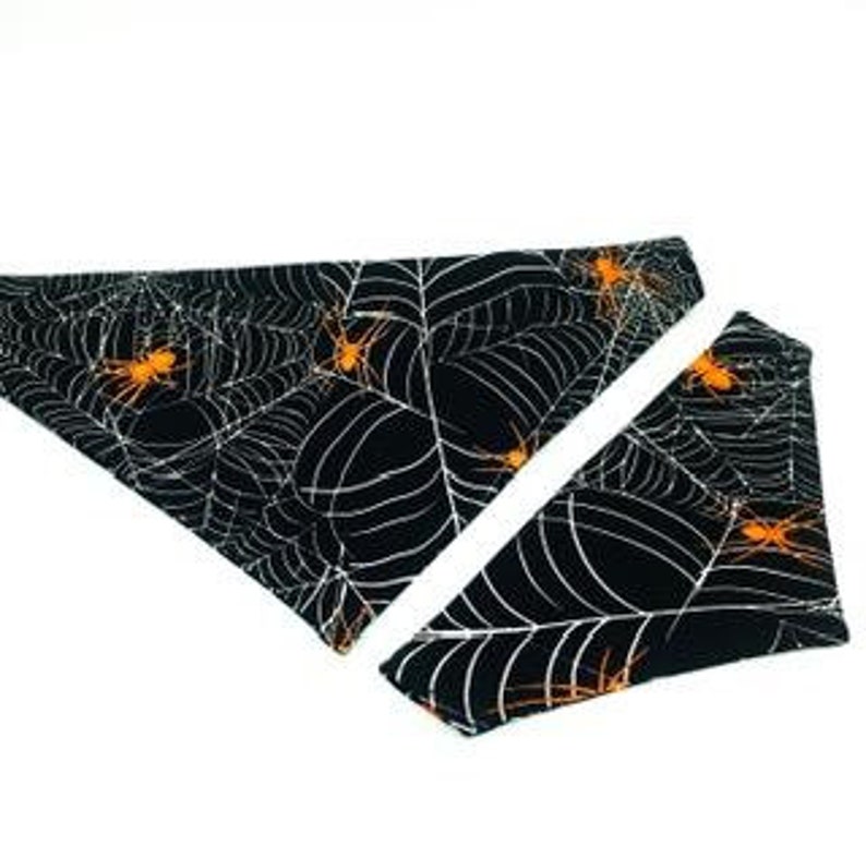 Op de afbeelding: Zwart-witte Halloween-bandana en bijpassend accessoire voor huisdieren. De stoffen items hebben een spinnenwebontwerp met oranje spinnen. De bandana en het accessoire worden op een witte achtergrond weergegeven.