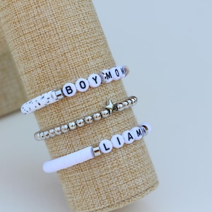 May include: Three beaded bracelets on display. One bracelet has white beads with black speckles. Another is silver with a star charm. The third bracelet has white letter beads spelling "LIAM". The bracelets are displayed on a tan fabric.