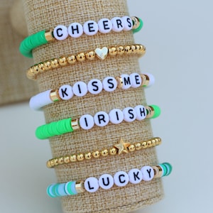 May include: Five bracelets with green and white beads and gold accents. The bracelets spell out "CHEERS", "KISS ME", "IRISH", "LUCKY", and one has a gold heart charm.