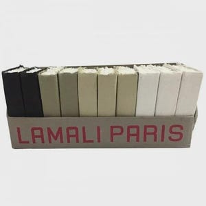 Pode incluir: Uma caixa de livros de cores sortidas com o texto "LAMALI PARIS" em vermelho. Os livros são em tons de preto, cinza, bege e branco. Os livros estão dispostos em fila.