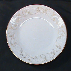 May include: White ceramic plate with a gold rim and decorative gold scrollwork around the edge. The plate has a slightly recessed center and is suitable for serving food. The plate is likely part of a dinnerware set.