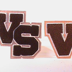May include: Two black felt patches with white stitching, one spelling "VS" and the other "V". The patches are on a white background.