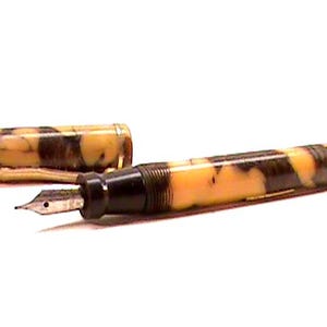 Puede incluir: Una pluma estilogr&aacute;fica vintage con un patr&oacute;n veteado en tonos amarillos y marrones. La pluma tiene una secci&oacute;n negra y un plum&iacute;n plateado. La tapa est&aacute; quitada, revelando el plum&iacute;n y el dise&ntilde;o de la pluma.