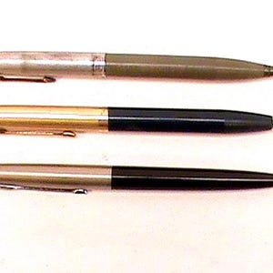 Könnte beinhalten: Drei Vintage-Stifte vor weißem Hintergrund. Der obere Stift hat eine silberne Kappe und einen grünen Körper. Der mittlere Stift hat eine goldene Kappe und einen schwarzen Körper. Der untere Stift hat eine silberne Kappe und einen schwarzen Körper.