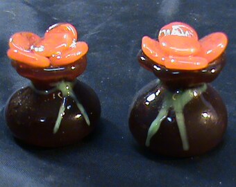 May include: Two small, dark brown glass vases with orange flower tops. The vases have a glossy finish and a white swirl design.