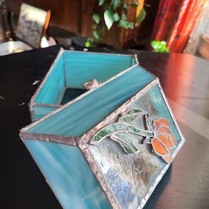 May include: A rectangular stained glass box featuring light blue marbled glass panels joined by silver-toned metal solder. The lid displays a decorative glass applique of a green stem with leaves and an orange flower bud attached to the surface.