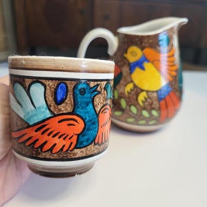 May include: A ceramic cup and pitcher set with a brown background and colorful bird designs. The cup features a blue bird with orange and blue wings. The pitcher has a white handle and a similar bird design with yellow, orange, and blue colors.