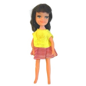 May include: A vintage doll with black hair and blue eyes. The doll is wearing a yellow textured top and a red and white striped skirt. The doll's legs are a light orange colour. A retro toy.