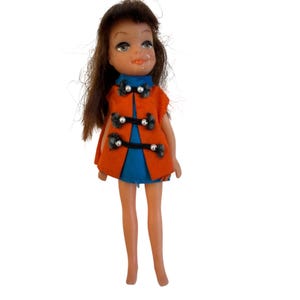 May include: A vintage doll with brown hair and a blue dress. The doll is wearing an orange vest with black and silver accents. The doll is standing on a white background.
