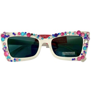 May include: White sunglasses with a square frame and rhinestones in various colors, including pink, blue, and purple. The sunglasses have a dark green lens and say "ULTRAVIOLET PROTECTION" on the right lens.