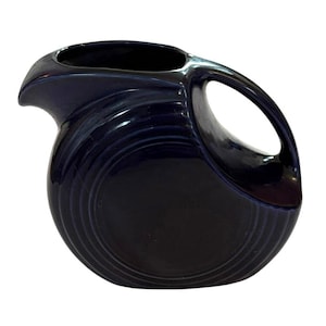 May include: A navy blue ceramic pitcher with a distinctive handle and spout. The pitcher has a rounded body with concentric circular ridges and a glossy finish. The handle is a large, open loop, and the spout is curved.