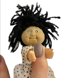 May include: A small doll with a round face, black yarn hair, and blue eyes. The doll is wearing a white dress with navy blue polka dots. The doll is being held by a person's hand.