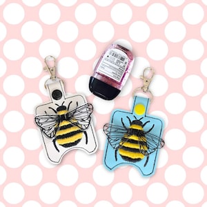 May include: Two bee-themed hand sanitizer holders with keychains. One is white with a yellow and black bee, the other is blue with a yellow and black bee. Both have a small yellow circle on the front.