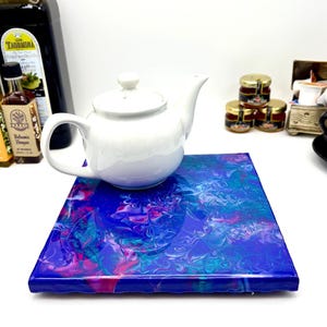 May include: A white ceramic teapot with a lid and handle sits atop a square coaster featuring a glossy, abstract acrylic pour design in vibrant shades of deep blue, purple, and turquoise with swirling patterns across the flat surface.