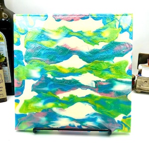 May include: Square abstract art piece with a fluid paint design. The artwork features a mix of colors, including blue, green, yellow, and pink, creating a unique and vibrant visual effect. The piece is displayed on a black metal stand.