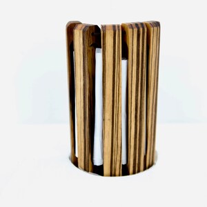 May include: A cylindrical wooden candle holder with vertical slats. The holder is made of light brown wood with dark brown stripes. White candles are visible inside the holder. The design is modern and minimalist.