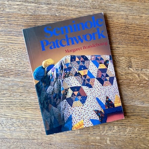 May include: A book titled "Seminole Patchwork" by Margaret Brandebourg. The cover features a colorful patchwork quilt design with blue, yellow, and white fabrics. Two balls of yarn in blue and yellow are resting on the book.