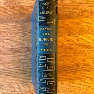 May include: A blue hardcover book titled "Prentice Hall Handbook for Writers, Second Edition." The spine features the authors' names and decorative geometric patterns in gold lettering. The book is on a wooden surface.