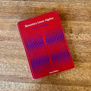 May include: A red book titled "Elementary Linear Algebra" by Paul C. Shields. The cover features a blue wavy design. The book is a second edition and rests on a wooden surface.