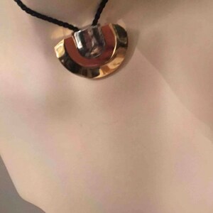 May include: A gold and silver pendant necklace with a black cord. The pendant is a geometric shape with a rounded top and a flat bottom.