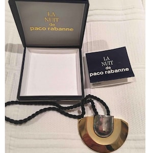May include: A gold and silver necklace with a large, round pendant. The pendant has a textured surface and is attached to a black cord. The necklace is in a box with the words "LA NUIT de paco rabanne" on it.