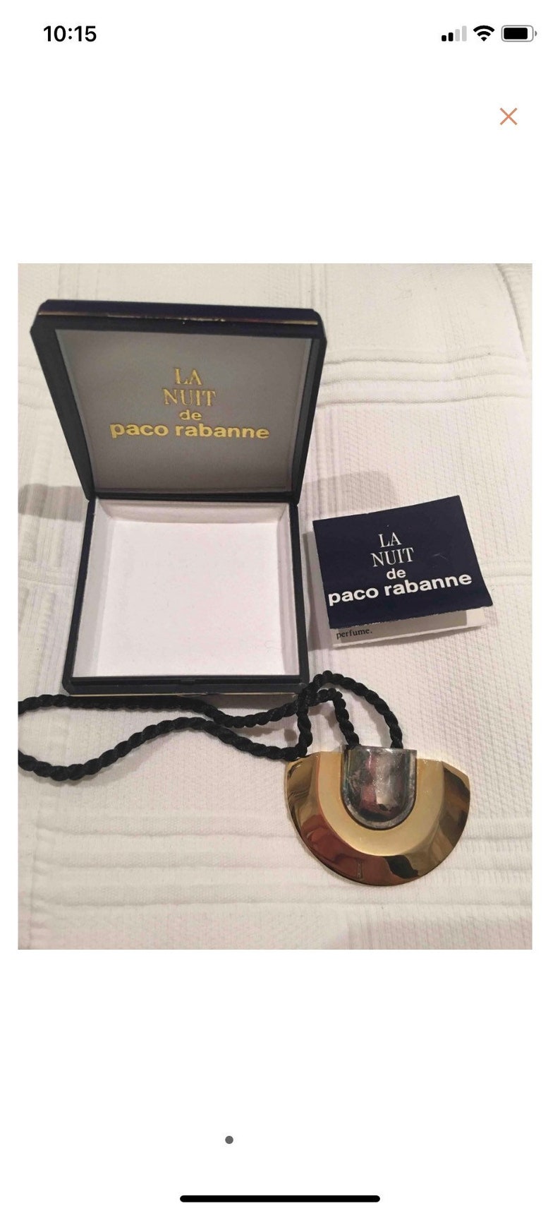 May include: A gold and silver necklace with a large, round pendant. The pendant has a textured surface and is attached to a black cord. The necklace is in a box with the words "LA NUIT de paco rabanne" on it.