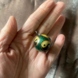 May include: A small, round, green and yellow pendant featuring a black and white yin-yang symbol. The pendant has a small loop at the top. The pendant is held in a hand.