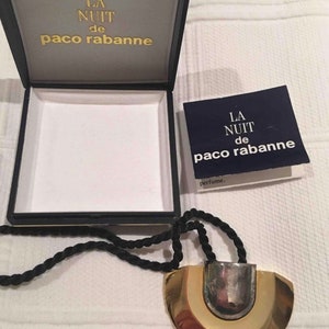 May include: A gold and silver necklace with a black cord. The pendant is a half-circle shape with a silver center and gold outer edges. The box is blue with gold lettering that reads "La Nuit de Paco Rabanne".