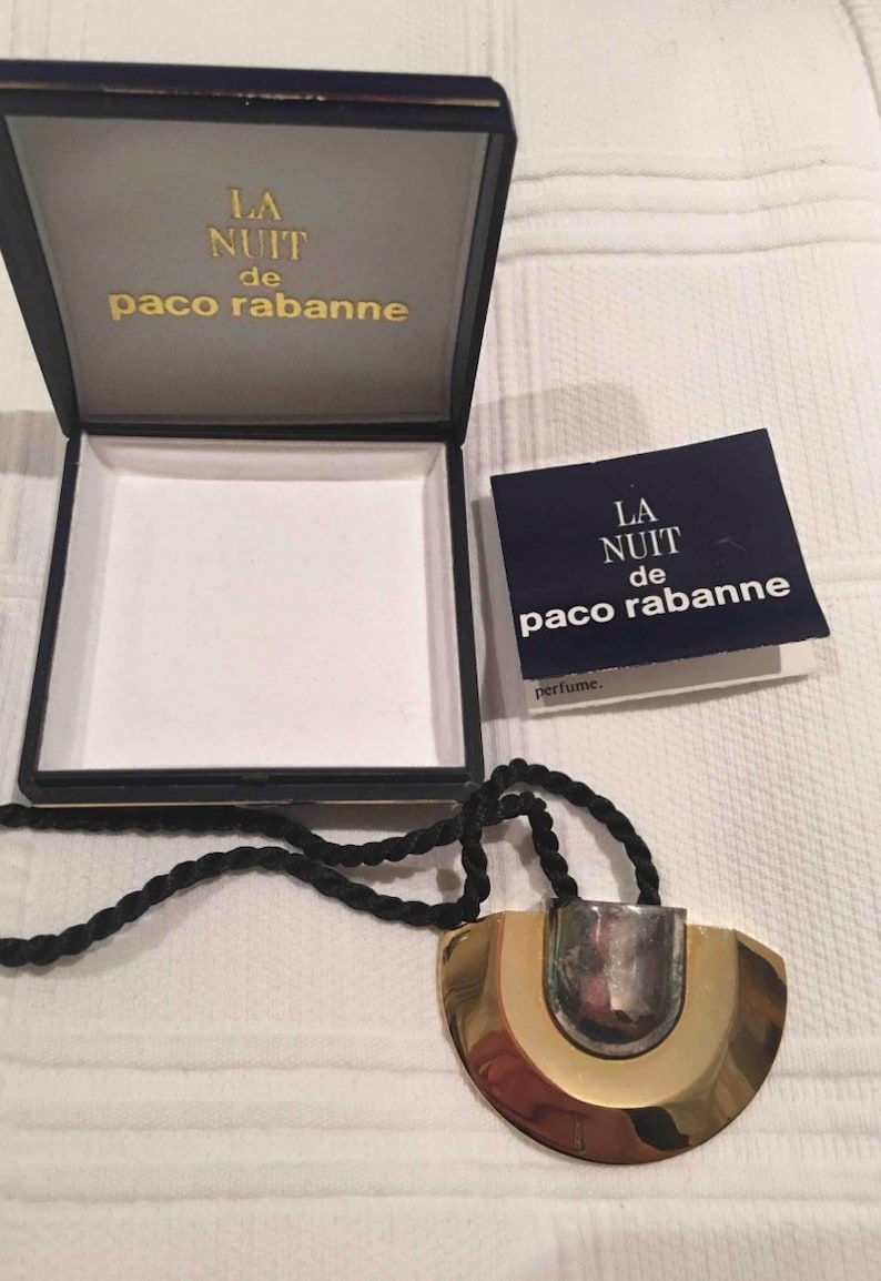 May include: A gold and silver necklace with a black cord. The pendant is a half-circle shape with a silver center and gold outer edges. The box is blue with gold lettering that reads "La Nuit de Paco Rabanne".