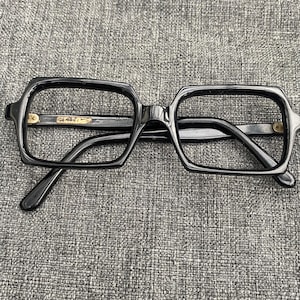 May include: Black square-shaped eyeglasses with gold accents on the arms. The glasses are on a gray textured surface.