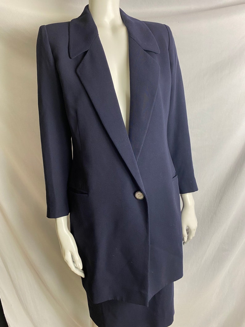 May include: A navy blue blazer and skirt suit with a single rhinestone button. The blazer has a long, fitted silhouette and a pointed collar.