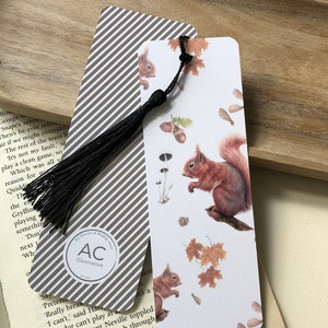 May include: Two decorative bookmarks. One features a squirrel illustration with autumn leaves and acorns, the other has a gray and white striped pattern. Both have a black tassel. The bookmarks are on a wooden tray.