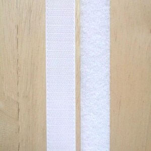 May include: Two strips of white hook and loop fastener tape, also known as Velcro, are shown against a light wood background.