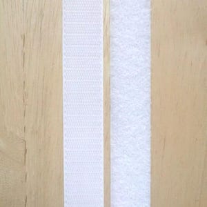 May include: Two strips of white hook and loop fastener tape, also known as Velcro, are shown against a light wood background.