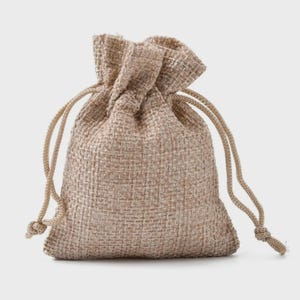 May include: A small, beige burlap drawstring bag. The bag is made of woven fabric with a textured appearance. The drawstring is a lighter beige colour and is knotted at the ends. The bag is closed at the top, with the drawstring pulled tight.