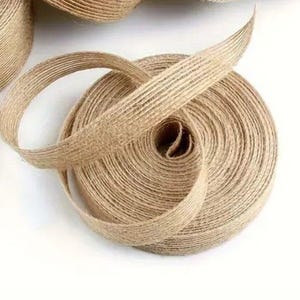 Może przedstawiać: Zbliżenie na rolkę naturalnej wstążki jutowej. Wstążka jest w jasnobrązowym kolorze i ma tkaną fakturę. Wstążka jest zwinięta w spiralę, z luźną pętlą wychodzącą ze środka.
