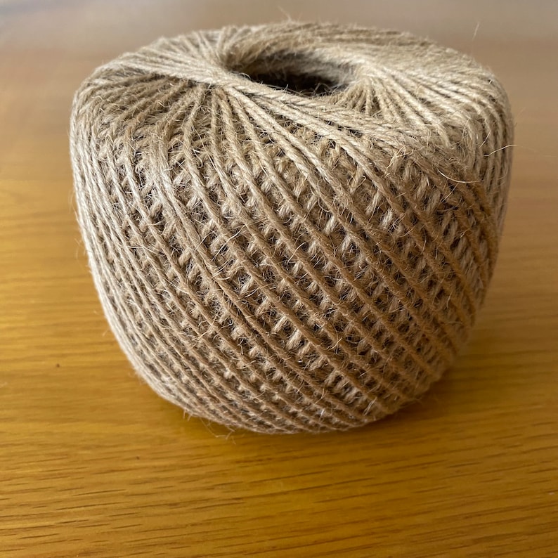 May include: A roll of natural brown twine, made from jute, is wound tightly in a circular shape. The twine is ideal for crafts, gardening, and packaging.