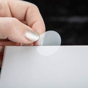May include: A clear, round sticker being peeled off of a white surface. The sticker is slightly translucent and has a white backing.
