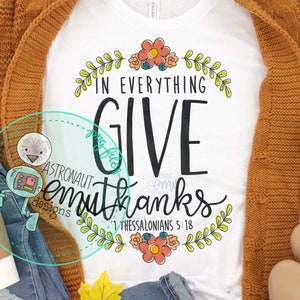 May include: A white t-shirt with a floral design and the text "In everything give thanks 1 Thessalonians 5:18". The shirt is styled with a brown cardigan, blue jeans, a coffee cup, and fall-themed accessories.