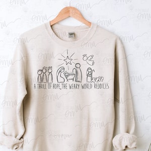 Puede incluir: Una sudadera beige con una ilustración negra de la escena de la natividad y el texto "A thrill of hope, the weary world rejoices".