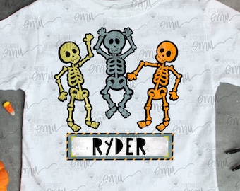 Op de afbeelding: Een wit T-shirt met een ontwerp van drie dansende skeletten. De skeletten zijn geel, blauw en oranje. De tekst "RYDER" is gedrukt op een zwart-geel bouwteken.