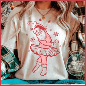 Könnte beinhalten: Hellbeige T-Shirt mit einer rosa Illustration von Weihnachtsmann im Tutu, der mit Schneeflocken tanzt. Das Shirt hat kurze Ärmel und einen Rundhalsausschnitt. Das Design ist eine skurrile Interpretation des Weihnachtsthemas.