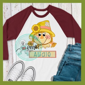 May include: A white and maroon baseball tee shirt with a cartoon scarecrow with a sunflower and the name "Audie" on a plaid banner.