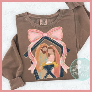May include: A brown Comfort Colors sweatshirt featuring a graphic of the Nativity scene. The design includes a pink bow, a blue and gold stable, and figures of Mary, Joseph, and baby Jesus. The sweatshirt has long sleeves.