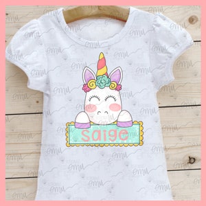 Op de afbeelding: Wit T-shirt met een eenhoorn afbeelding die een bord vasthoudt met de tekst "Saige". De eenhoorn heeft een regenbooghoorn, roze en paarse bloemen en gesloten ogen. Het bord is lichtblauw met een gele rand.